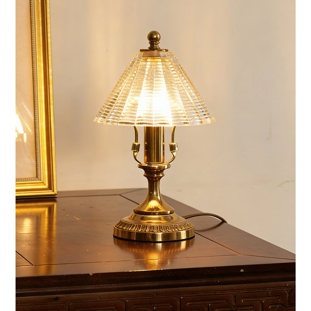 Front view of this Clérachic lamp lit on a polished wood table, the brass stem and ribbed clear glass shade catching the light.