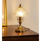 Front view of this Clérachic lamp lit on a polished wood table, the brass stem and ribbed clear glass shade catching the light.