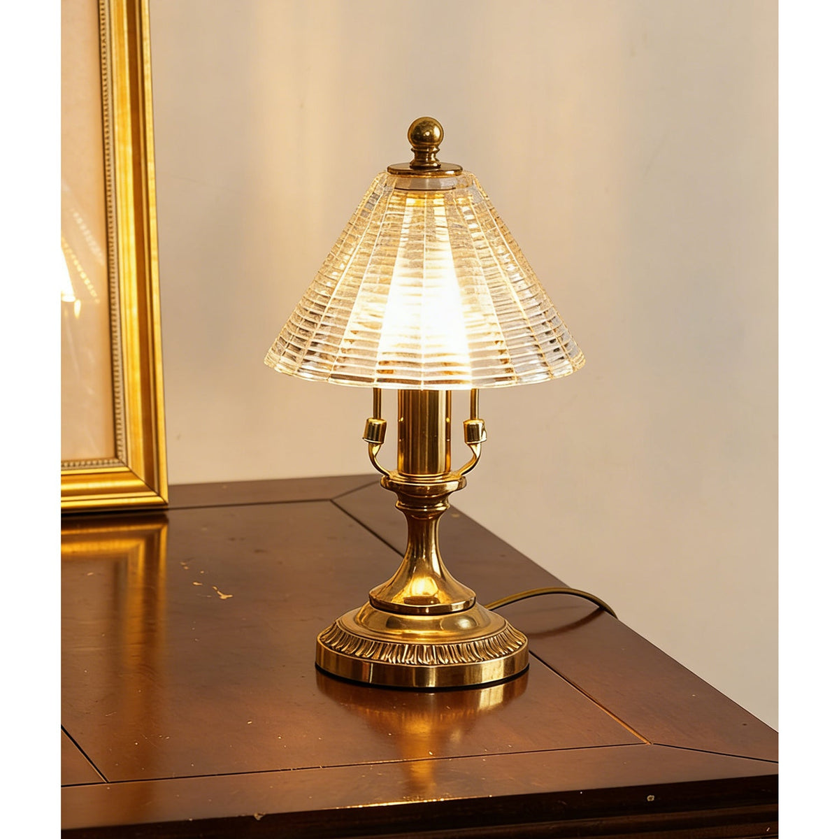 Angled view of the lit Clérachic table lamp, its antique brass base reflected in the polished wood tabletop beside a gilt frame.
