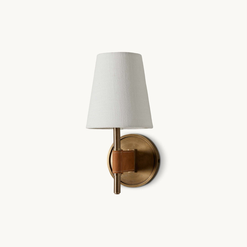 Front view of this Clérachic wall light on a plain white backdrop, with a white fabric shade, an antique brass arm and a tan strap at the backplate.