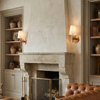 A Clérachic wall light glowing beside a limewashed stone fireplace, with shelves of pottery and a tan leather sofa in the room.