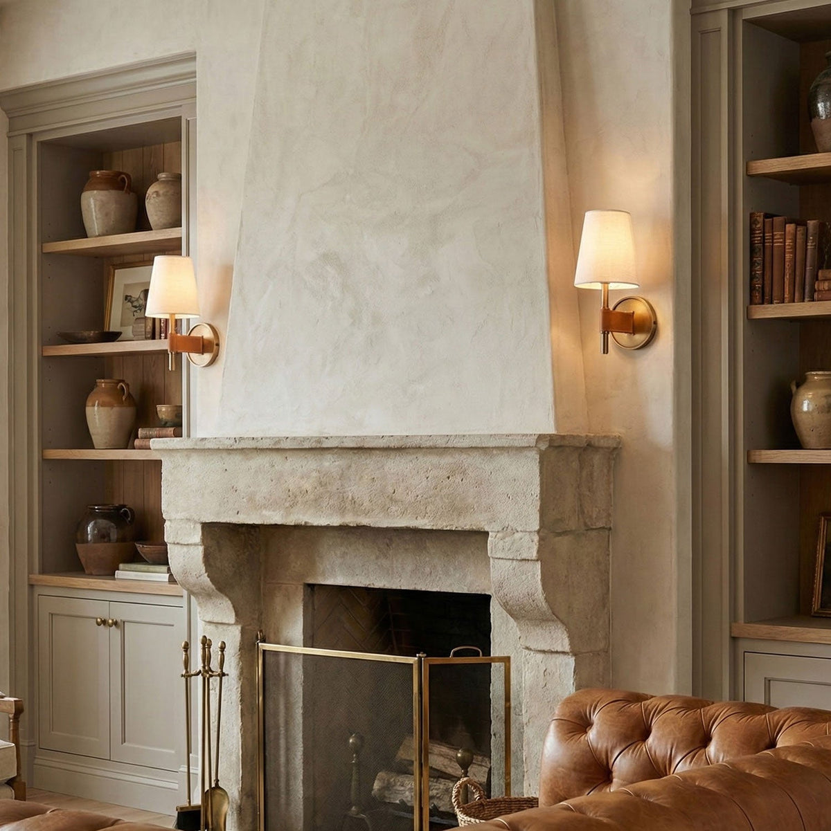 A Clérachic wall light glowing beside a limewashed stone fireplace, with shelves of pottery and a tan leather sofa in the room.
