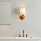 Single Clérachic wall light with a lit white shade above a basin with brass taps, on a pale grey wall.