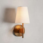 Close, straight-on view of a single Clérachic wall light, with a lit linen-look shade and a round antique brass backplate with a tan strap.