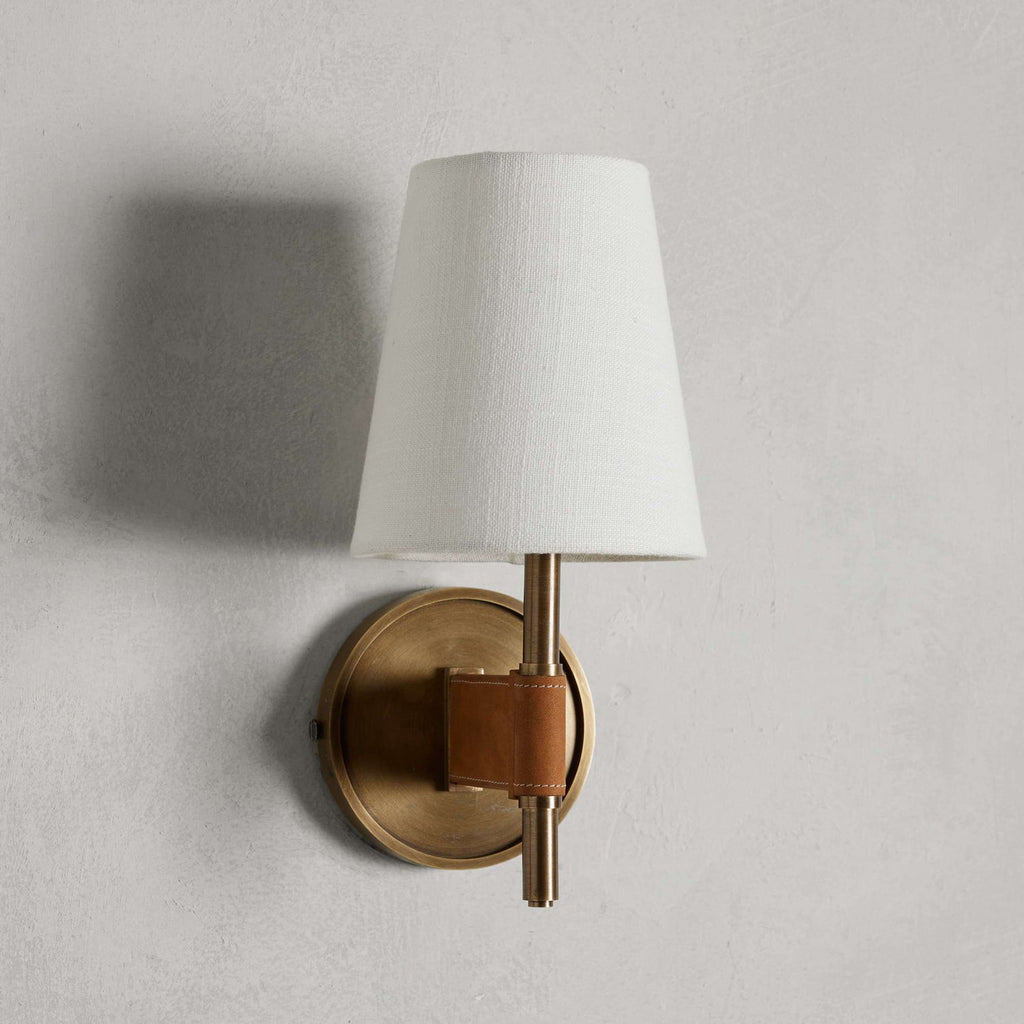 Unlit white tapered shade on this Clérachic wall light, held on a short brass stem above a round antique brass backplate on a grey wall.