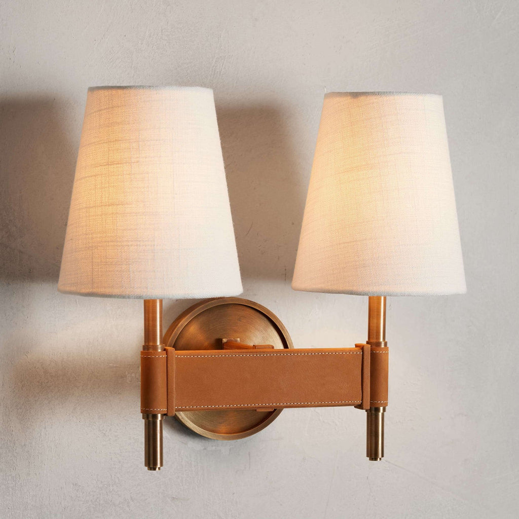 Double Clérachic wall light lit warmly, two linen-look shades on brass stems joined by a wide tan stitched strap across a round backplate.