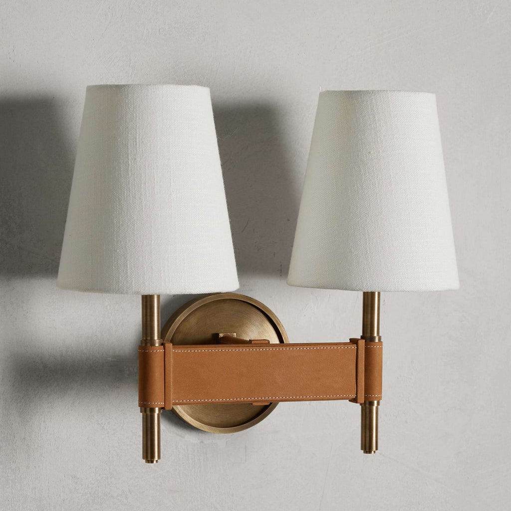 Unlit double Clérachic wall light on a grey wall, two white tapered shades above a tan stitched crossbar and a small round brass backplate.
