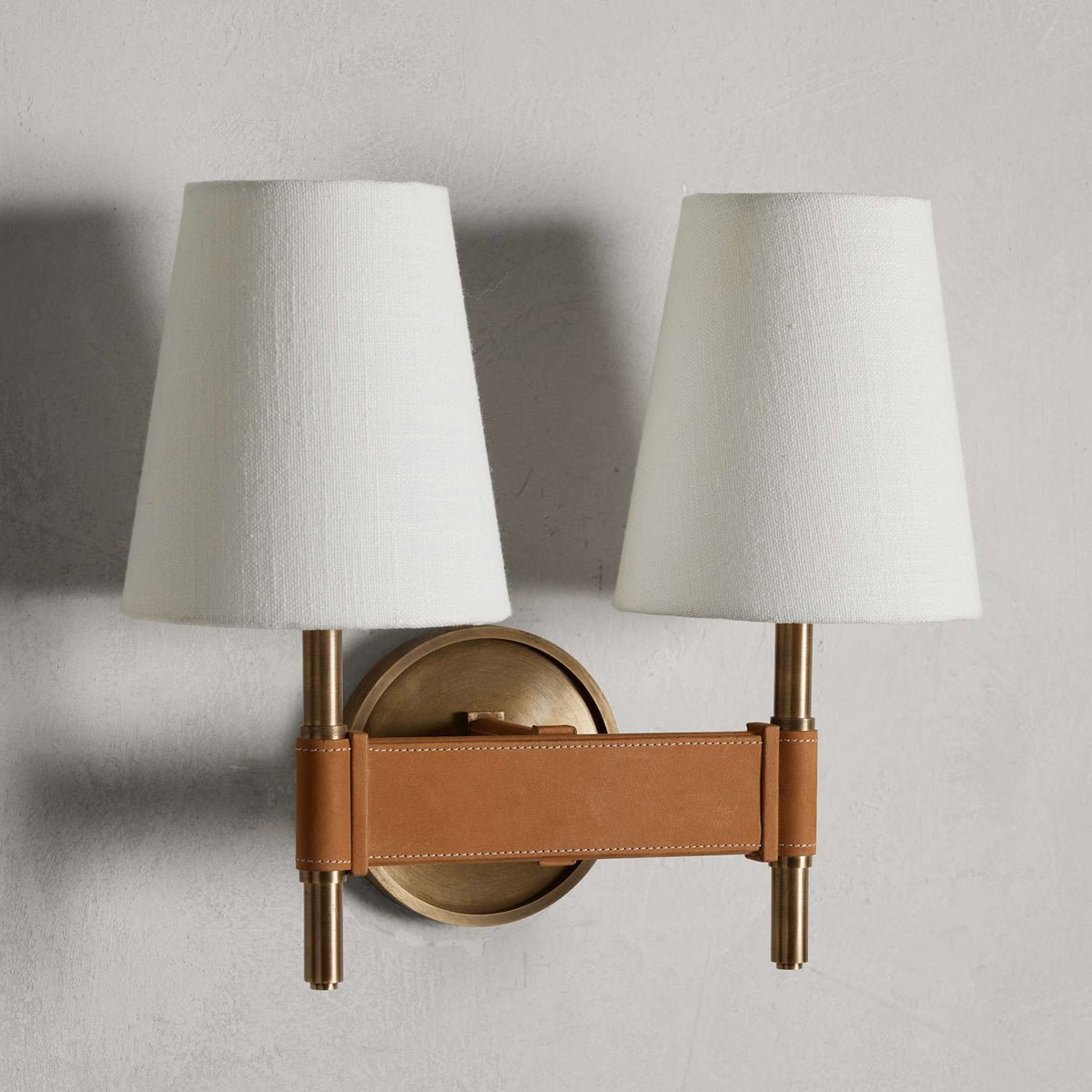Unlit double Clérachic wall light on a grey wall, two white tapered shades above a tan stitched crossbar and a small round brass backplate.