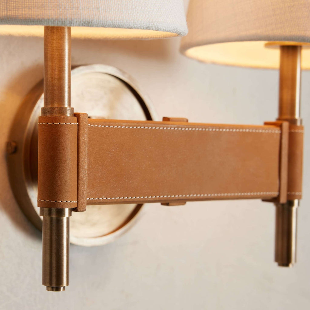 Close view of the stitched tan crossbar joining two brass stems on this Clérachic double wall light, with the shade rims at the top.