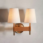 Double Clérachic wall light lit on a grey wall, with two cream shades above a tan stitched crossbar and a round brass backplate.