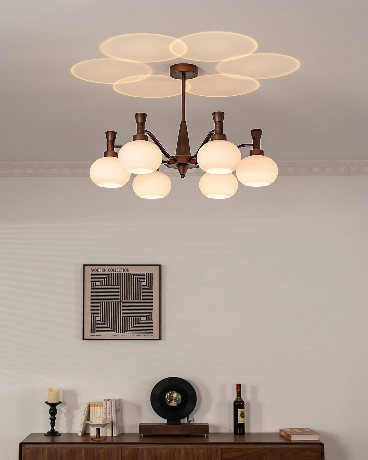 A Clérachic antique bronze chandelier lit above a wall print and a wooden sideboard holding a record player and a bottle.