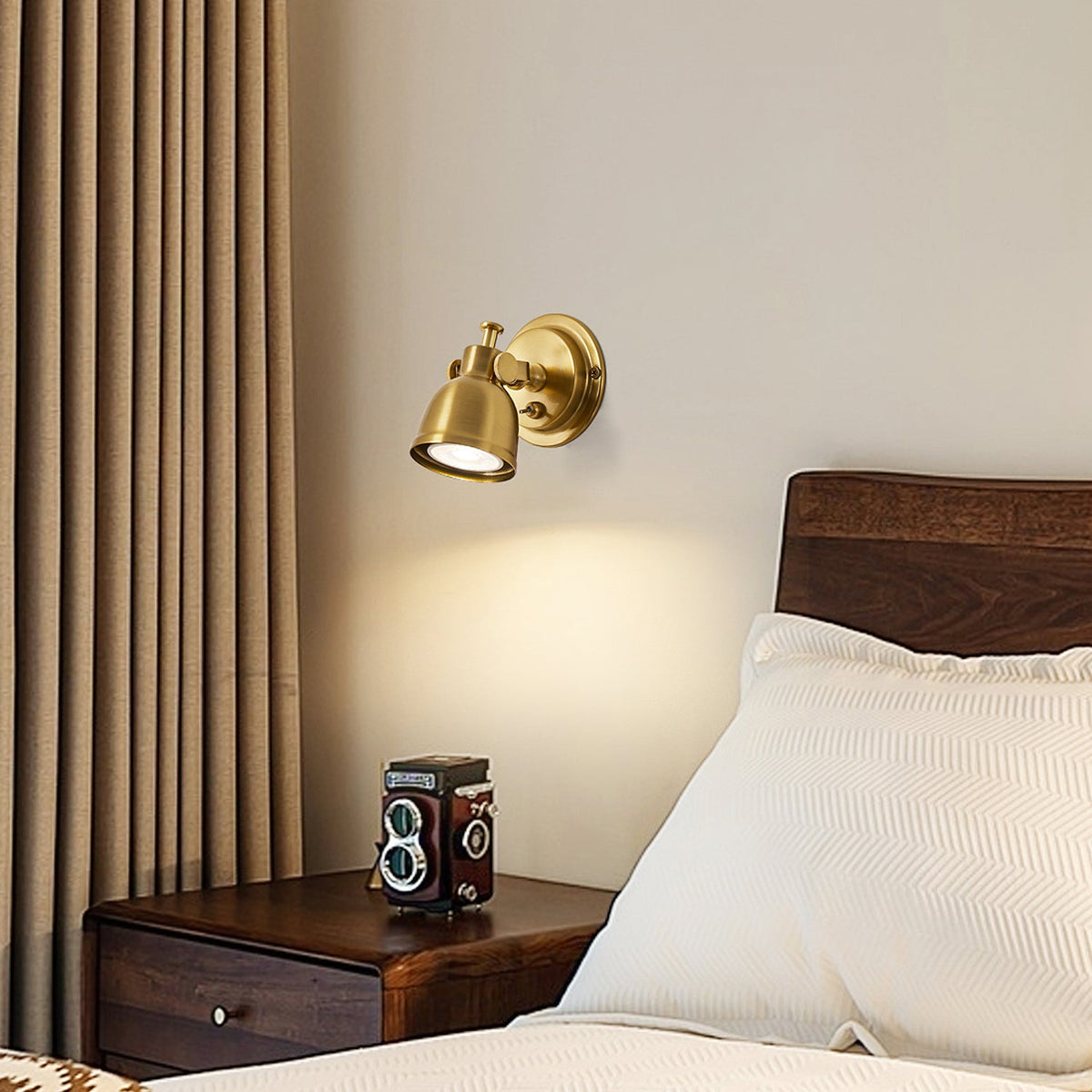 A Clérachic wall light glowing warm beside a walnut bed, with a cream pillow and curtain and a vintage camera on the bedside table.