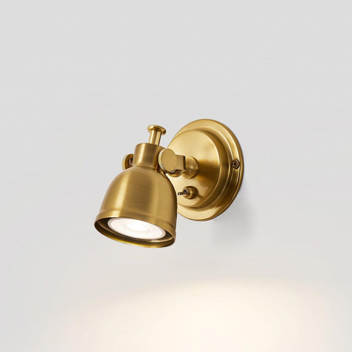 Side view of the brass dome shade of this Clérachic wall light, lit, with a soft fan of light on the pale grey wall below.