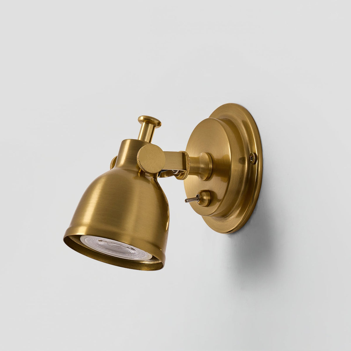 Three-quarter view of this Clérachic wall light in antique gold, with the swivel arm and toggle switch visible against a light grey wall.