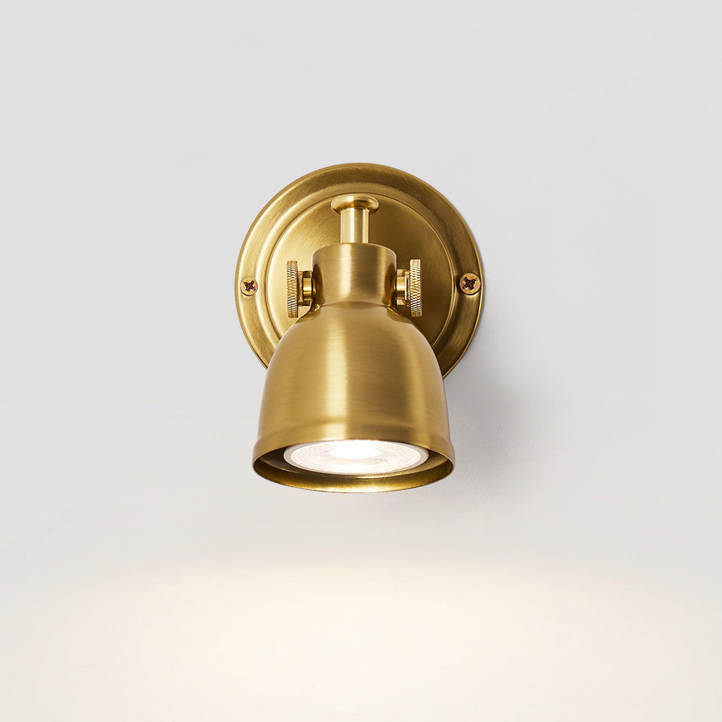 Front view of this Clérachic wall light, its round backplate behind a brushed gold dome with the bulb lit and a glow below on grey.