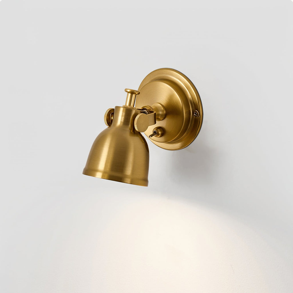 Clérachic wall light angled downwards, the brass shade tilted on its swivel joint with light spreading across the grey wall beneath.