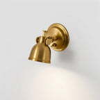 Clérachic wall light angled downwards, the brass shade tilted on its swivel joint with light spreading across the grey wall beneath.