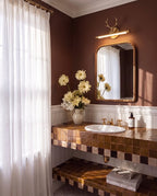 Clérachic brass antler wall light glowing above a framed mirror in a brown-walled washroom with a tiled vanity and sheer curtain.
