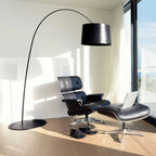 Black Clérachic arc floor lamp reaching over a leather lounge chair and footstool beside a sunlit window, on a light oak floor.