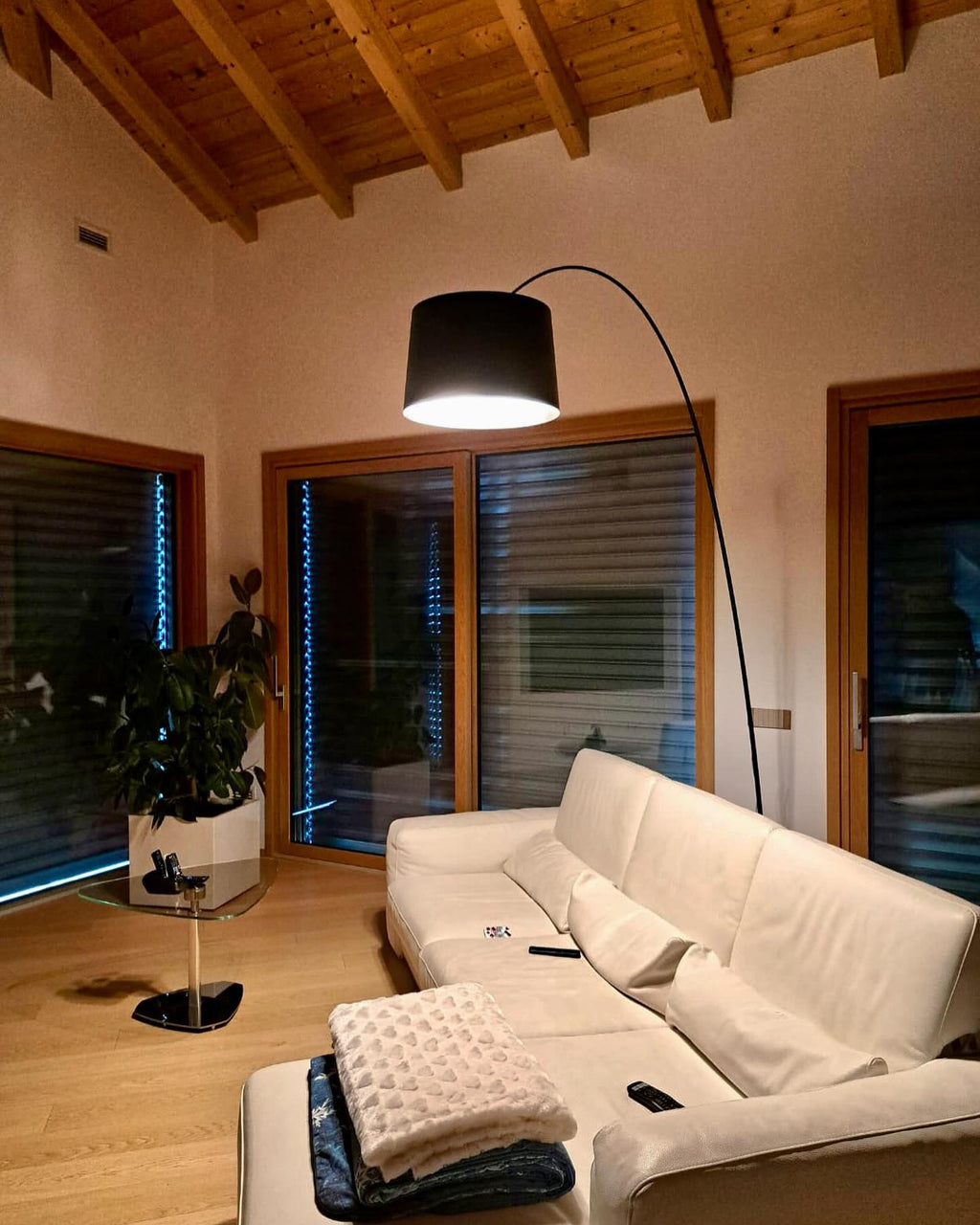 Black Clérachic arc floor lamp lit in a timber-ceilinged room, its arm arching over a cream sofa and a leafy plant.
