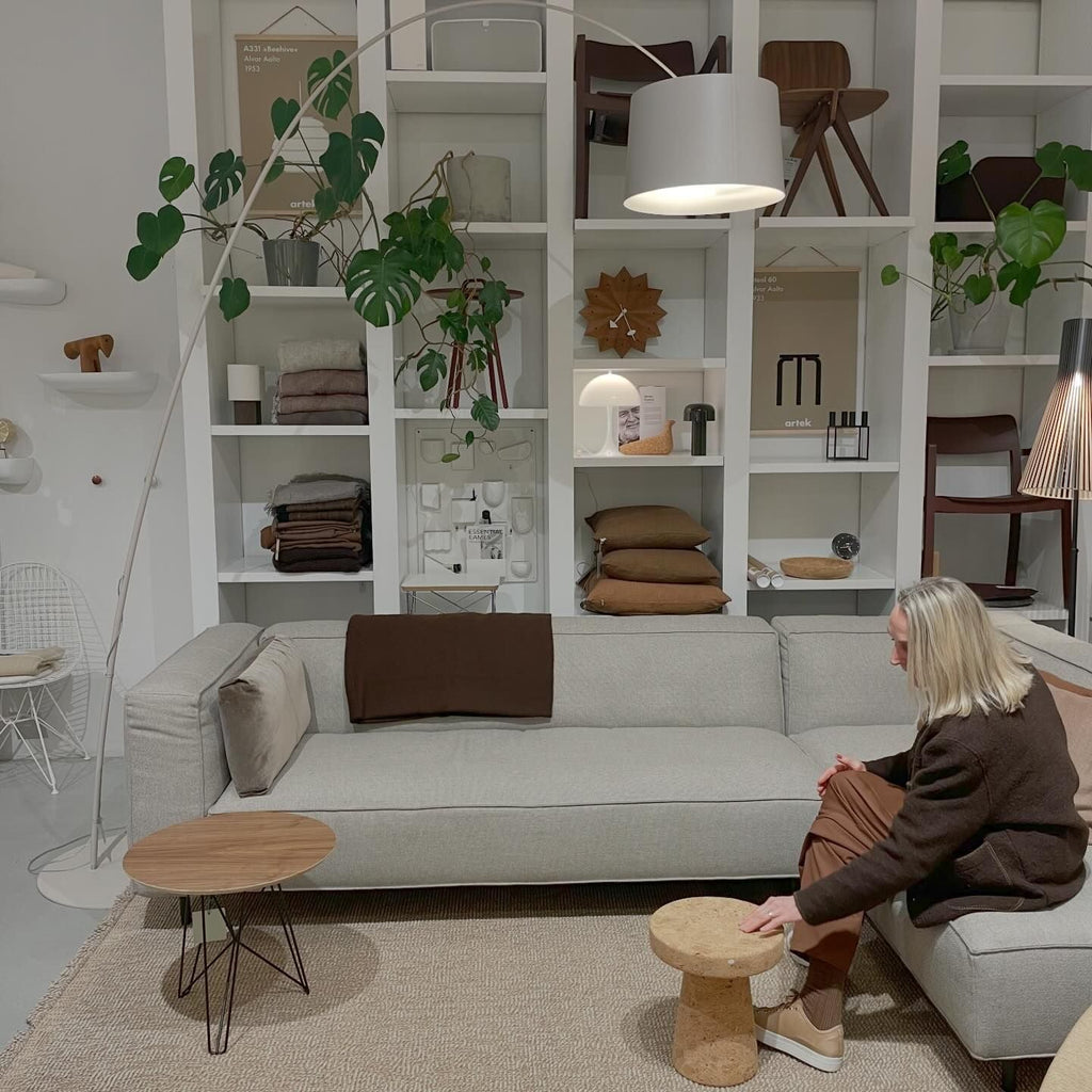 White Clérachic arc floor lamp with a pale drum shade, arching over a grey sofa in front of a white shelving wall.