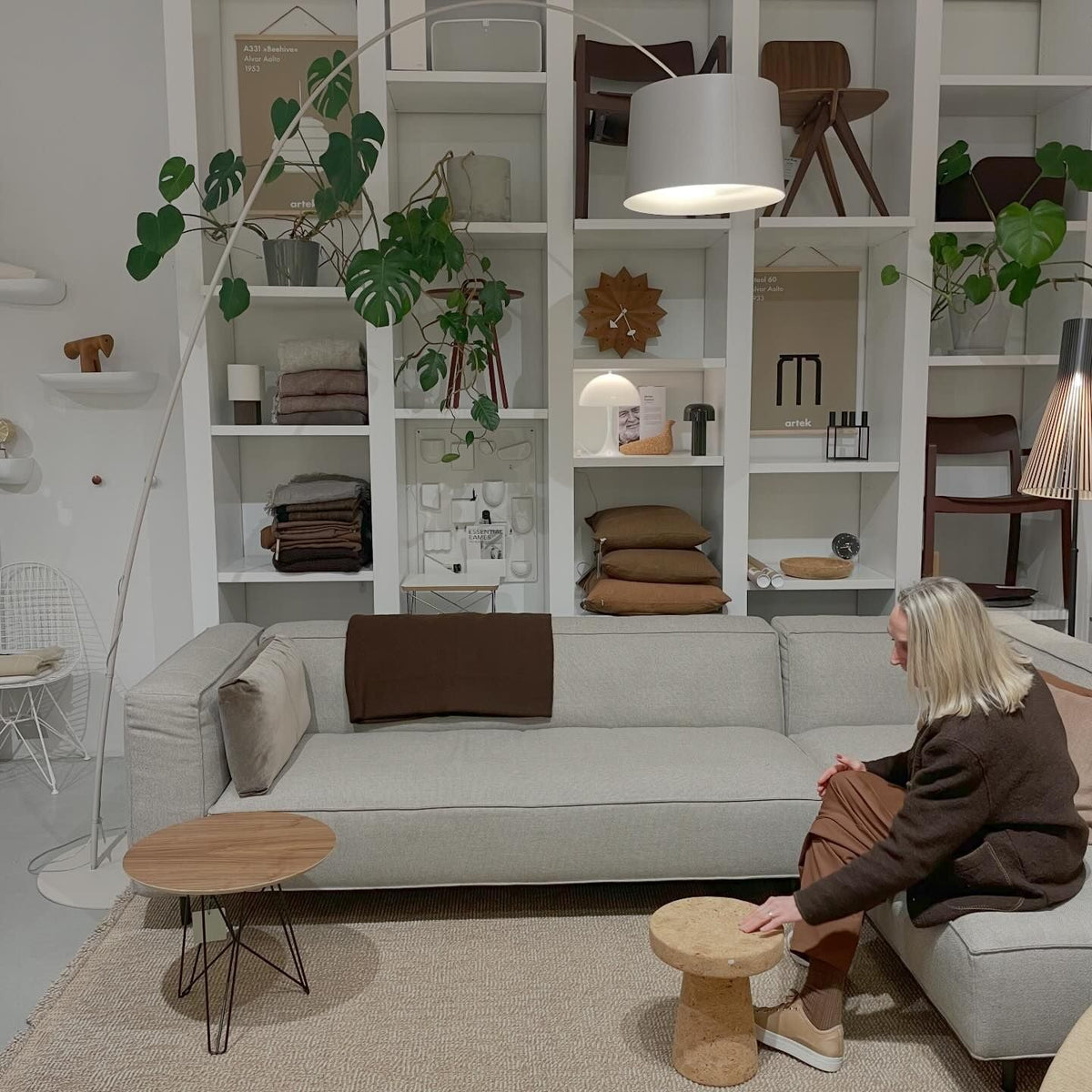 White Clérachic arc floor lamp with a pale drum shade, arching over a grey sofa in front of a white shelving wall.