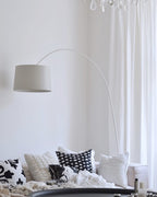 White Clérachic arc floor lamp with a grey-white shade arching over a sofa piled with black and white cushions.