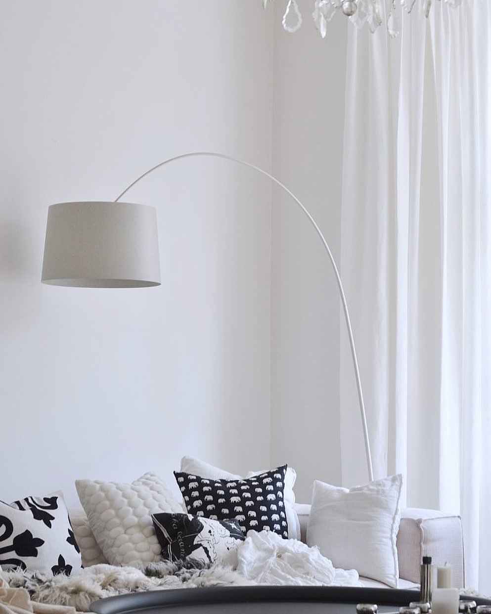 White Clérachic arc floor lamp with a grey-white shade arching over a sofa piled with black and white cushions.
