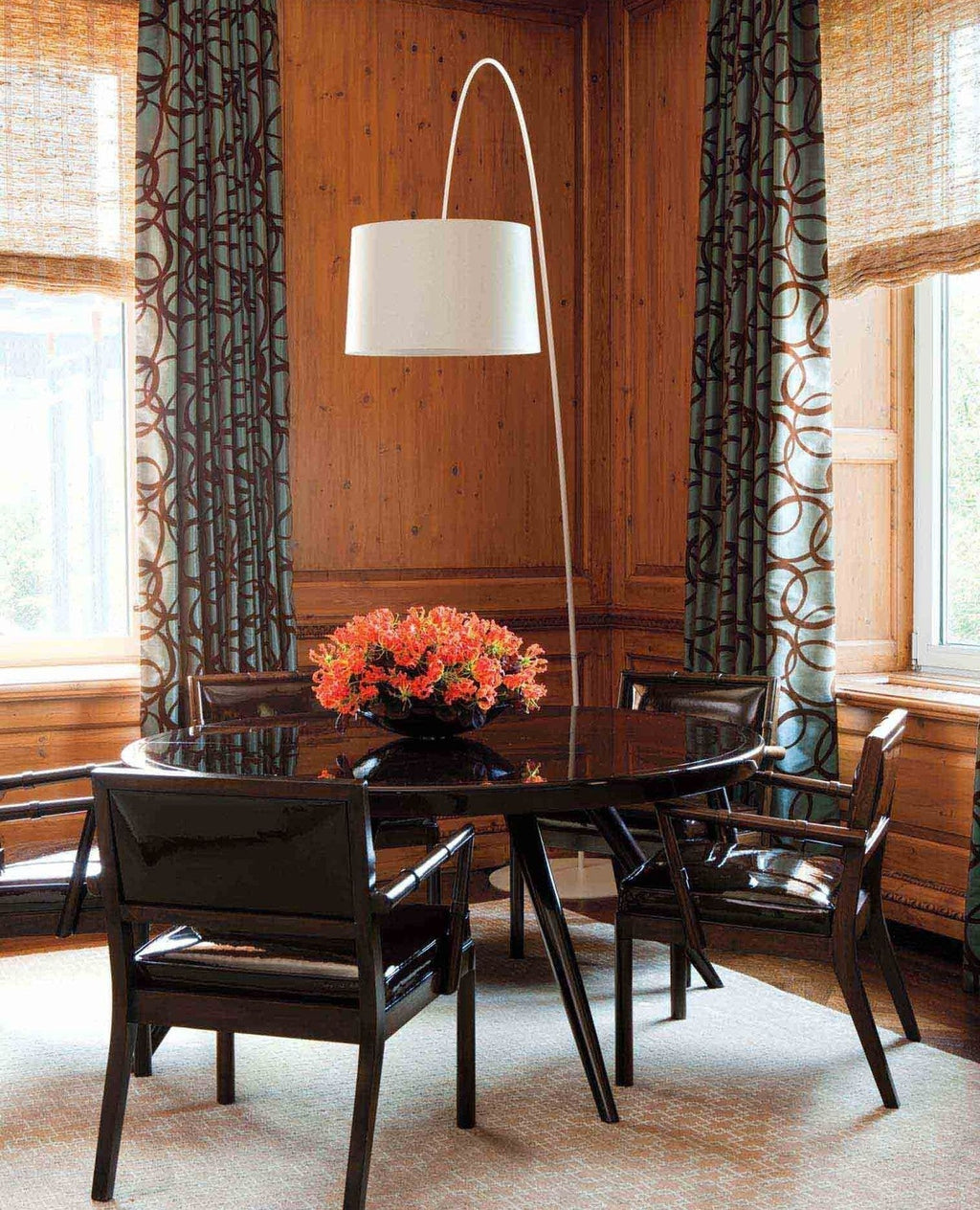 White Clérachic arc floor lamp rising behind a round dark dining table, with patterned curtains and warm wood panelling.