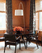 White Clérachic arc floor lamp rising behind a round dark dining table, with patterned curtains and warm wood panelling.