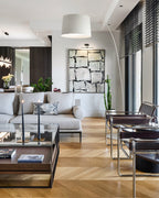 White Clérachic arc floor lamp over a living room with a grey sofa, a glass coffee table and a herringbone wood floor.