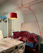 Red Clérachic arc floor lamp with a red drum shade, its curved arm reaching over a tufted velvet sofa and a laid table.