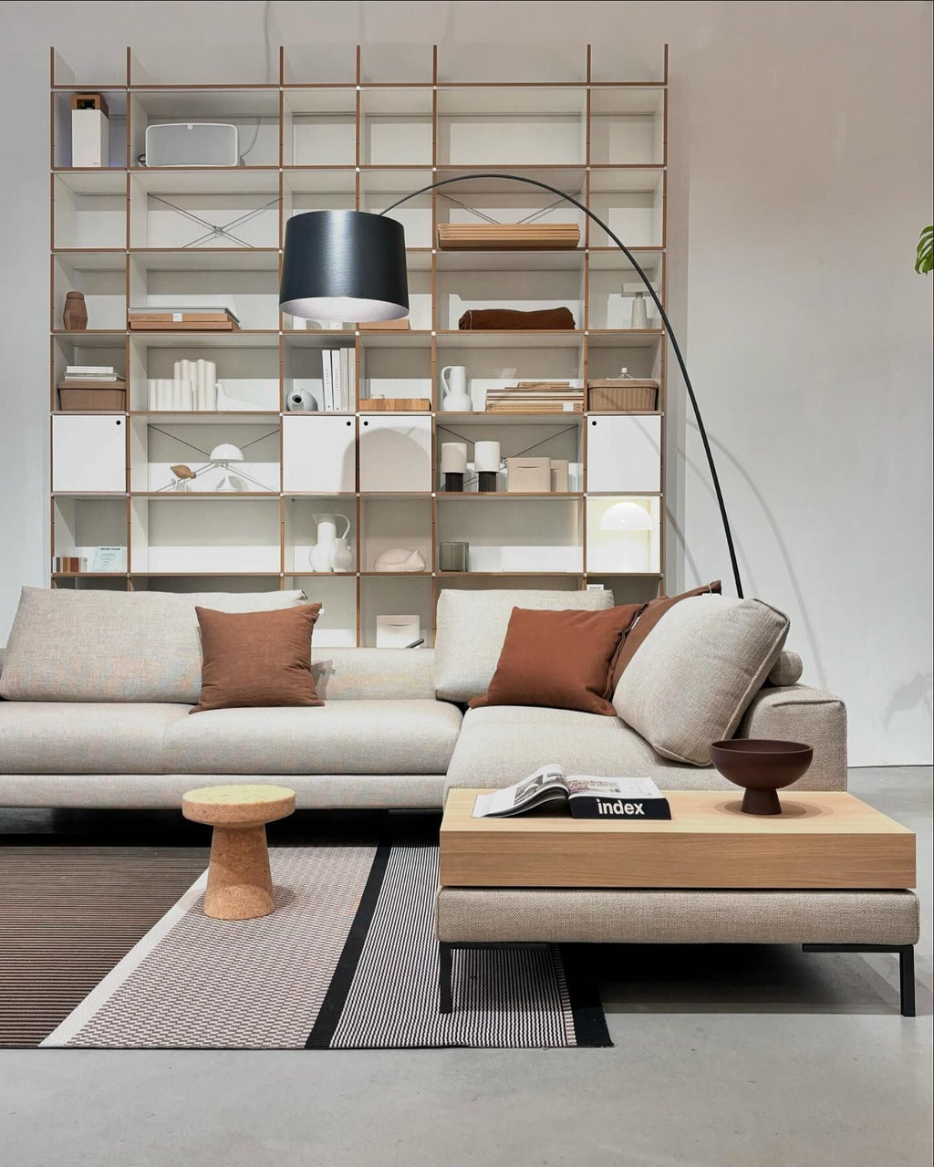 Black Clérachic arc floor lamp arching over a cream sectional sofa in front of an open white shelving unit.