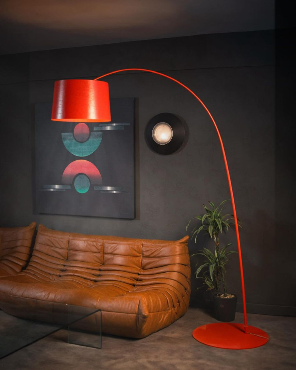 Red Clérachic arc floor lamp with a round red base beside a tan leather sofa and a dark grey wall.