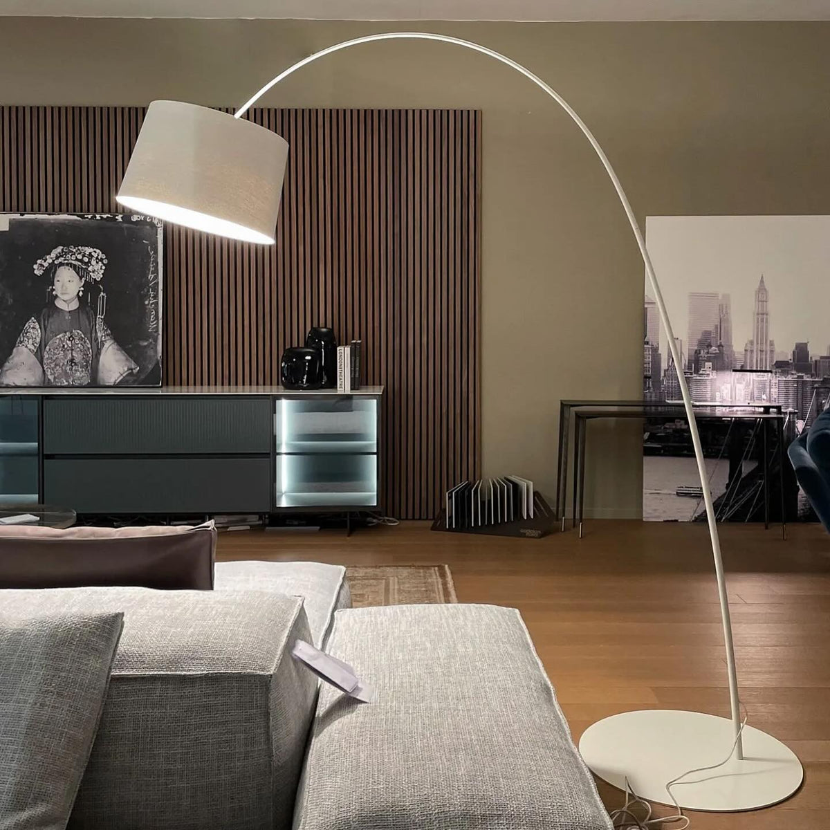 White Clérachic arc floor lamp with a white drum shade and base, arching over a grey modular sofa.