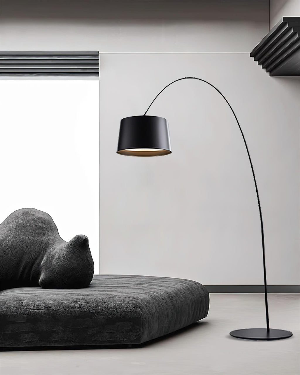 Black Clérachic arc floor lamp arching over a low charcoal daybed in a white room, with the round base on a grey floor.