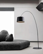 Black Clérachic arc floor lamp arching over a low charcoal daybed in a white room, with the round base on a grey floor.