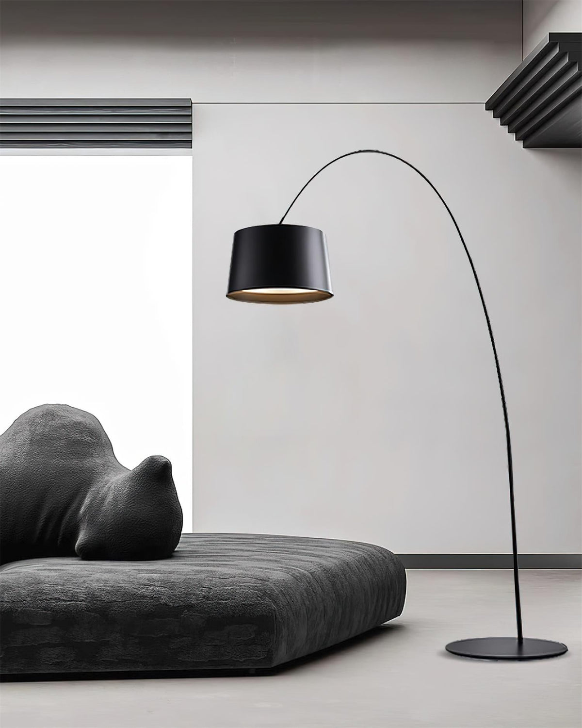 Black Clérachic arc floor lamp arching over a low charcoal daybed in a white room, with the round base on a grey floor.