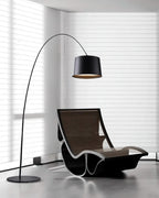 Black Clérachic arc floor lamp beside a black rocking lounger in a corner with white horizontal blinds.