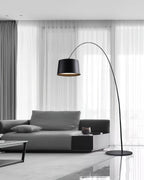 Black Clérachic arc floor lamp reaching over a grey sofa in front of sheer curtains in a bright living room.