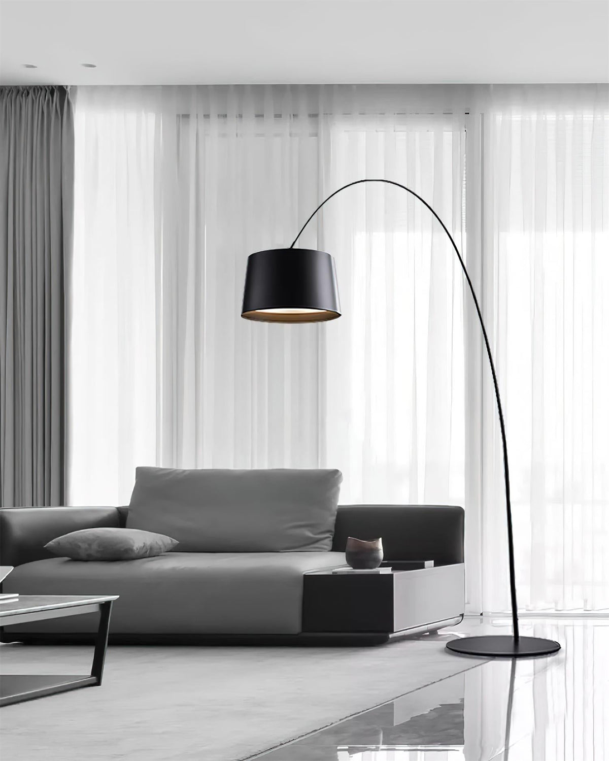 Black Clérachic arc floor lamp reaching over a grey sofa in front of sheer curtains in a bright living room.