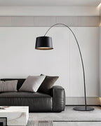 Black Clérachic arc floor lamp beside a black leather sofa with grey cushions, a marble table and a pale grey wall behind.