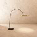 Clérachic arc floor lamp with a thin black arm and a gold ring light, on a round black base against a limewash wall.