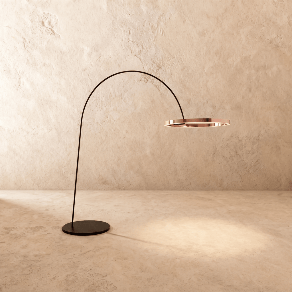 Clérachic arc floor lamp with a polished rose gold ring on a black arm, casting a soft pool of light on the floor.