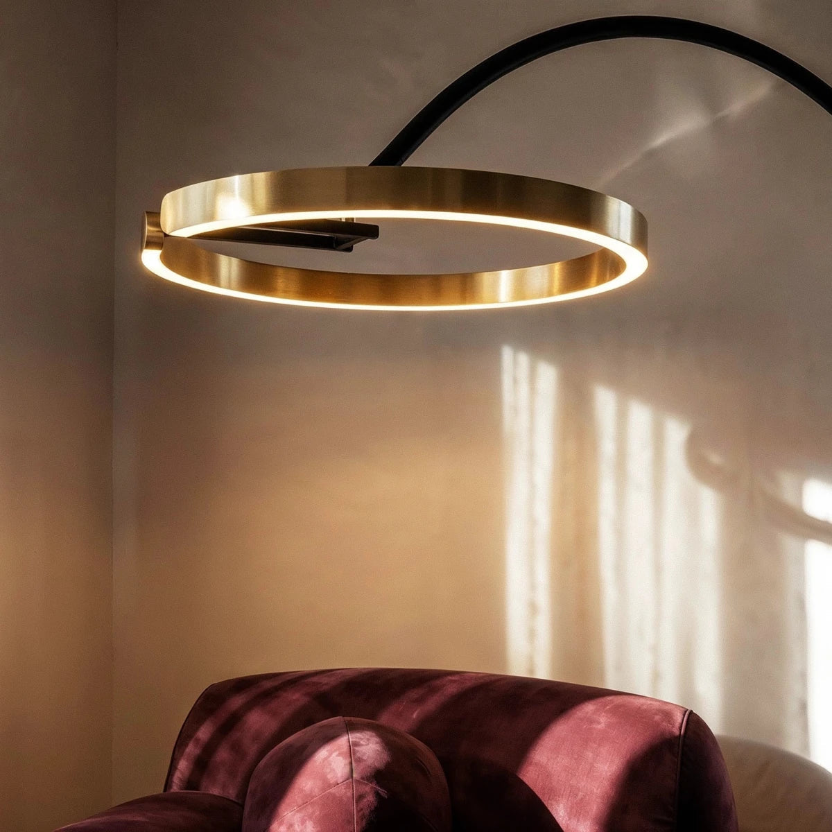 Close view of the Clérachic brushed gold ring light, joined to the black arm and glowing warmly beside a velvet armchair.