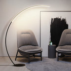 Clérachic black arc floor lamp curving over two grey fabric lounge chairs, the lit band along the top of its bow washing warm light up the wall beside a black abstract canvas.