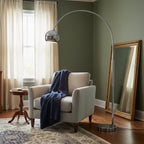 A Clérachic chrome arc lamp curves over a grey armchair with a navy throw, beside a gilt floor mirror and a sage green wall.