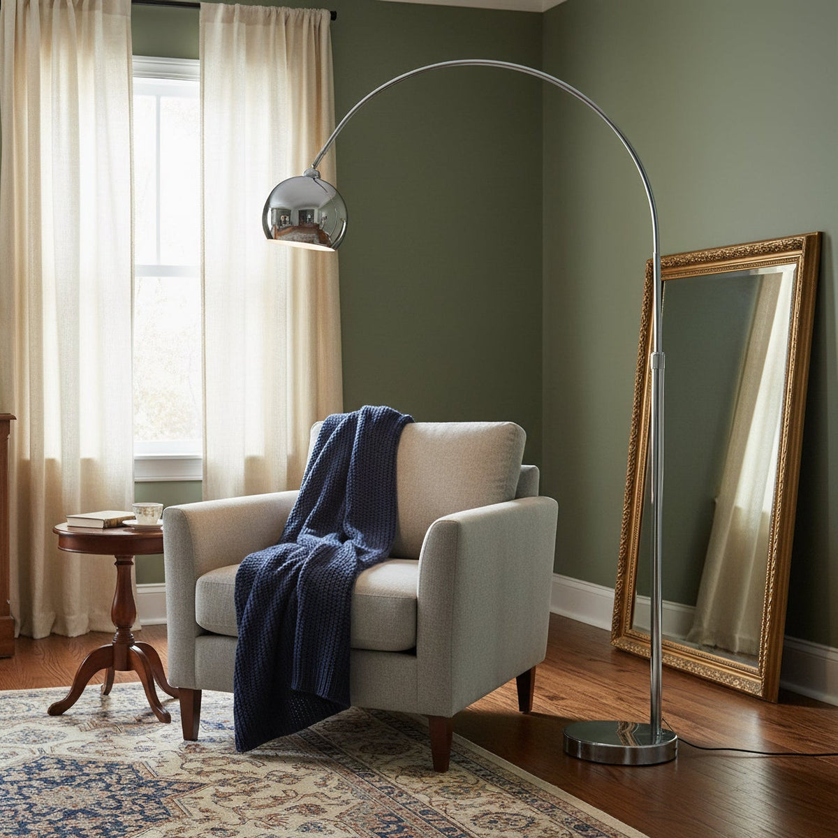 A Clérachic chrome arc lamp curves over a grey armchair with a navy throw, beside a gilt floor mirror and a sage green wall.