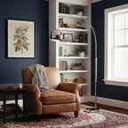 Chrome arc floor lamp from Clérachic arching over a tan leather armchair, with built-in shelves and a navy wall behind.