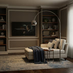 This Clérachic arc lamp leans over a cream chaise with a dark throw, in a den with a wall-mounted screen and built-in shelves.